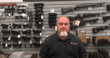 Bob Bailey in gun store discussing AIM Weapons Grade CLP Videos with various firearms on display.